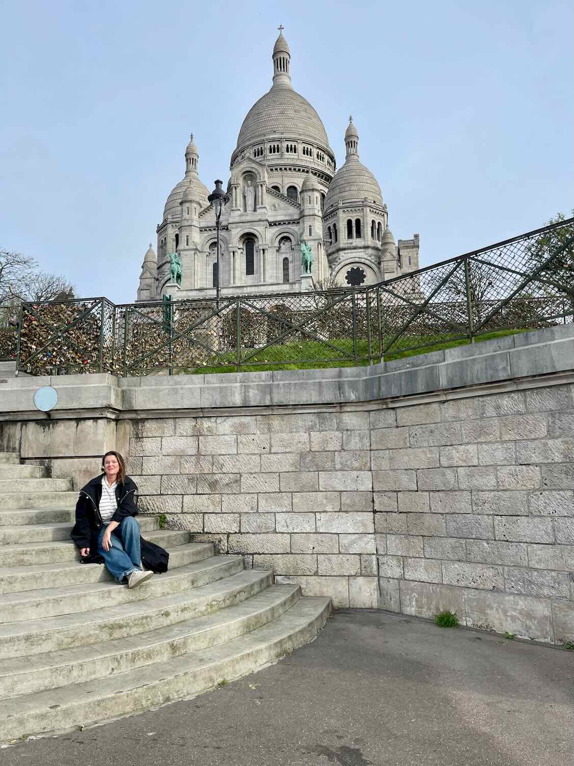A solo female traveler sits on the steps in front of the majestic Sacré-Cœur Basilica in Paris. The intricate white architecture of the basilica rises impressively behind her, under a clear sky.