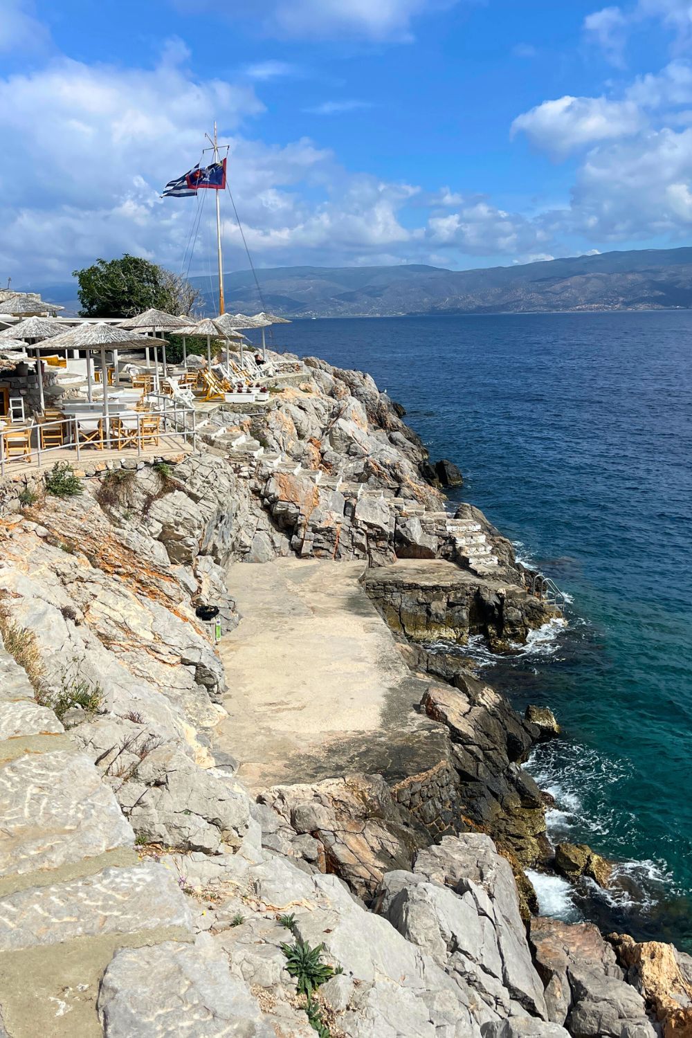 Seaside leisure at its best in Hydra, with a rocky beach adorned with straw umbrellas and wooden loungers overlooking the azure sea and distant mountains.
