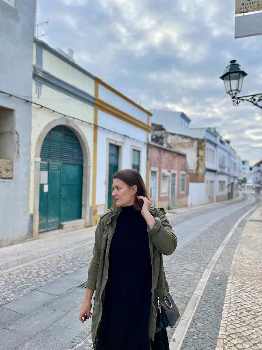A woman in a casual black dress and green jacket walking along a cobblestone street in Faro, with historical buildings lining the road and a street lamp hanging above.