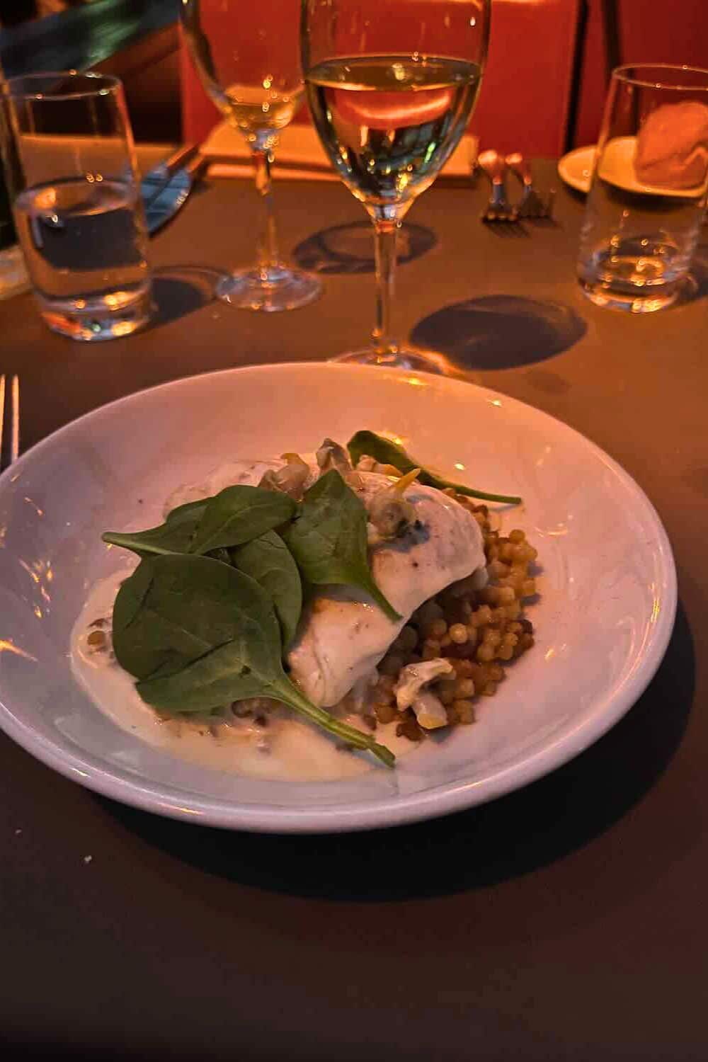 A plate of fish served over a bed of lentils and topped with fresh spinach, presented on a white plate in an intimate dining setting with ambient lighting and glasses of water and wine.