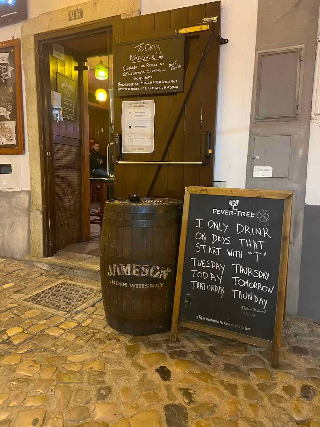 An entrance to a bar in Tavira with a blackboard sign humorously listing days starting with 'T' as drinking days, next to a Jameson Irish Whiskey barrel.