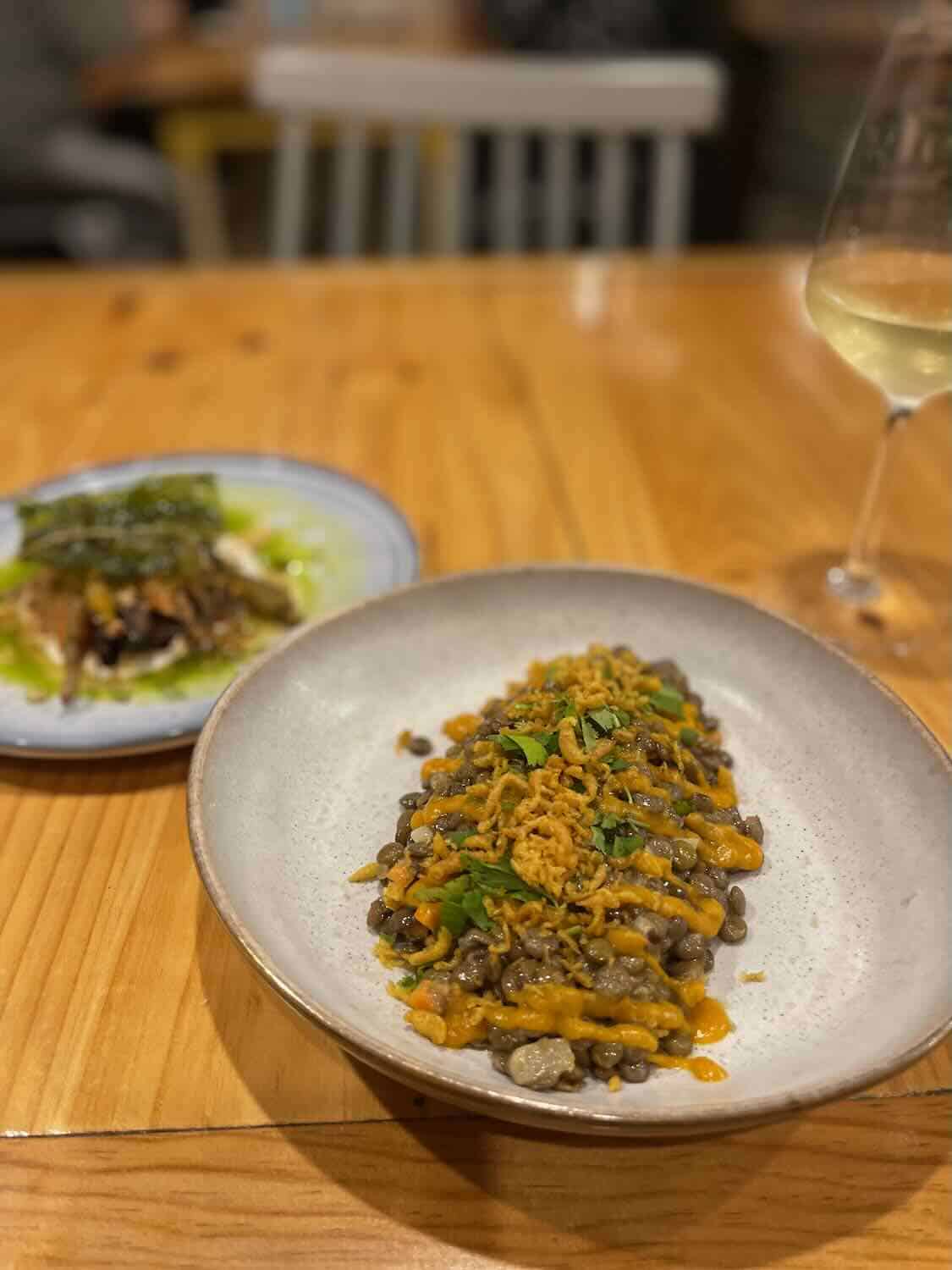 A gourmet plate in the foreground with lentils and vibrant orange sauce topped with crispy onions, accompanied by a glass of white wine and an out-of-focus dish in the background, presenting a cozy dining atmosphere.