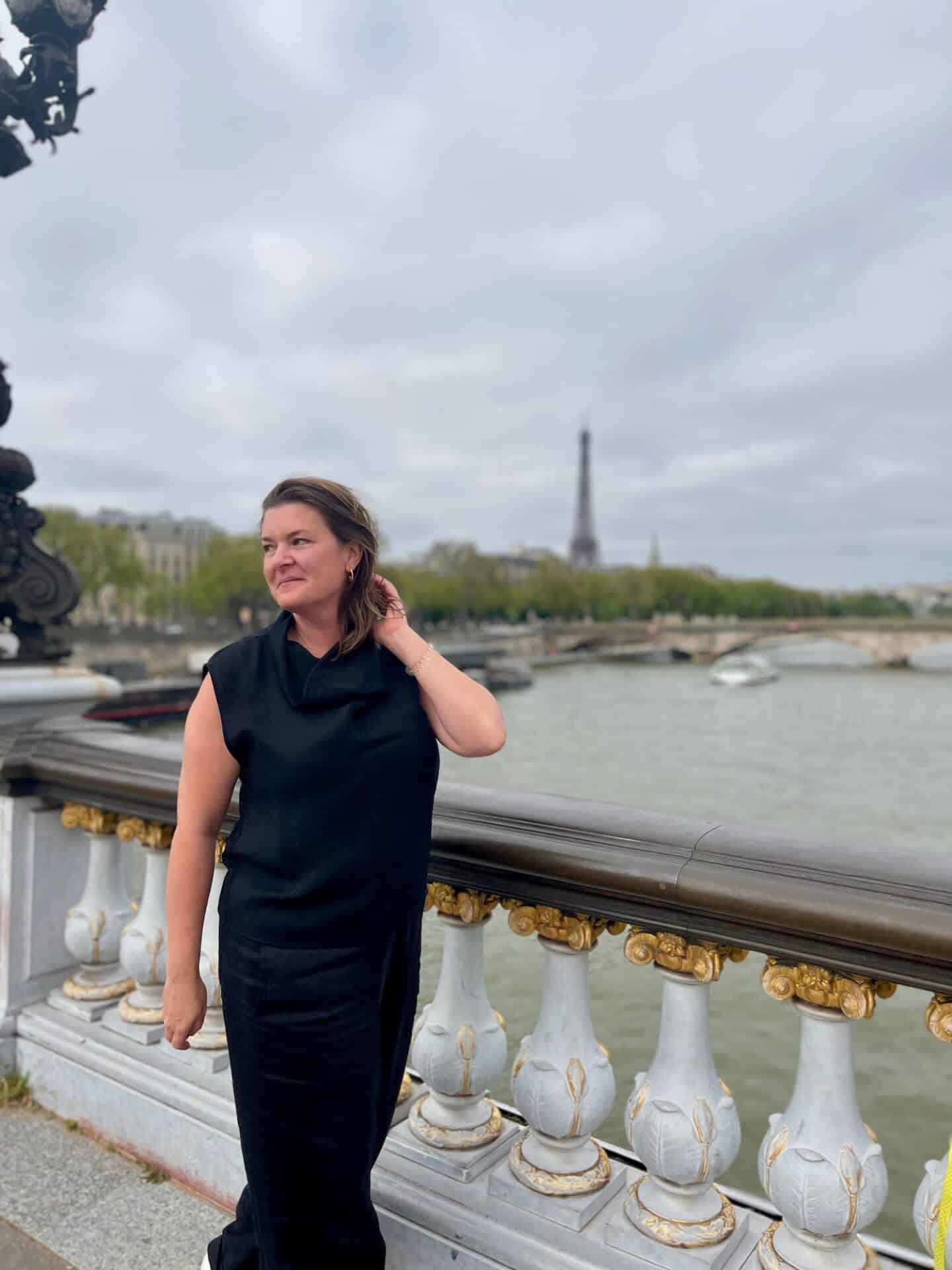 A woman in elegant black attire poses thoughtfully on a Parisian bridge, with the iconic silhouette of the Eiffel Tower in the background. The soft light of an overcast sky gives the scene a serene ambiance, reflecting the contemplative solitude of solo travel.