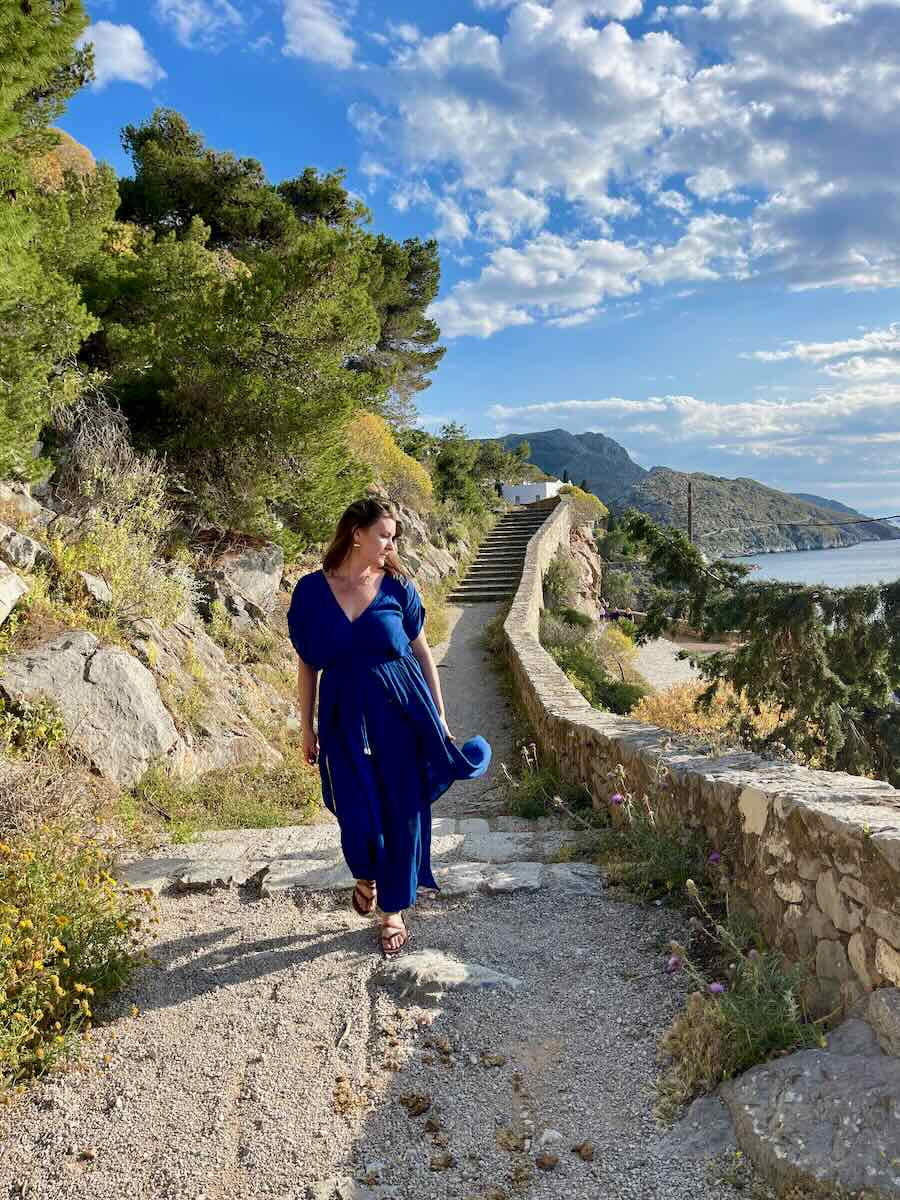 A woman in a blue dress walks along a scenic path on Hydra, with the Mediterranean Sea and rugged landscape as a backdrop, reflecting a moment of tranquil exploration on the Greek island.