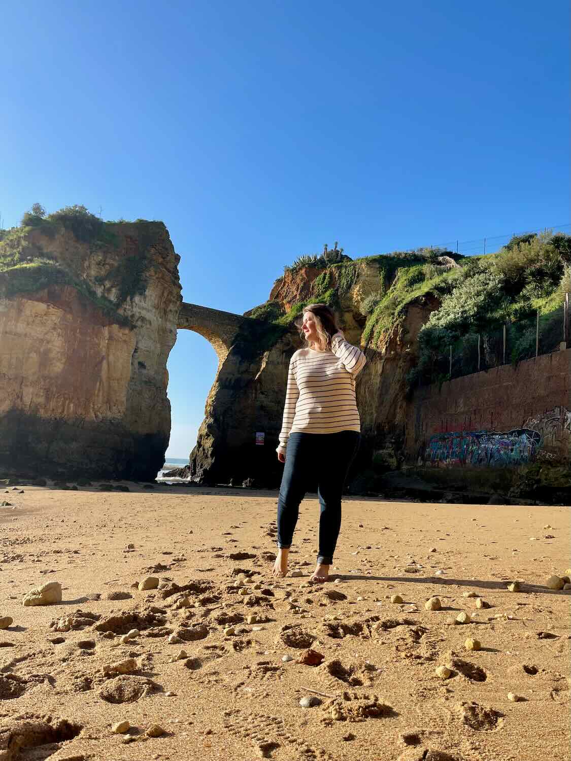 A woman stands barefoot on a sandy beach, with rocky cliffs and a stone arch bridge in the background. The sky is clear and blue, and there are scattered footprints and rocks on the sand