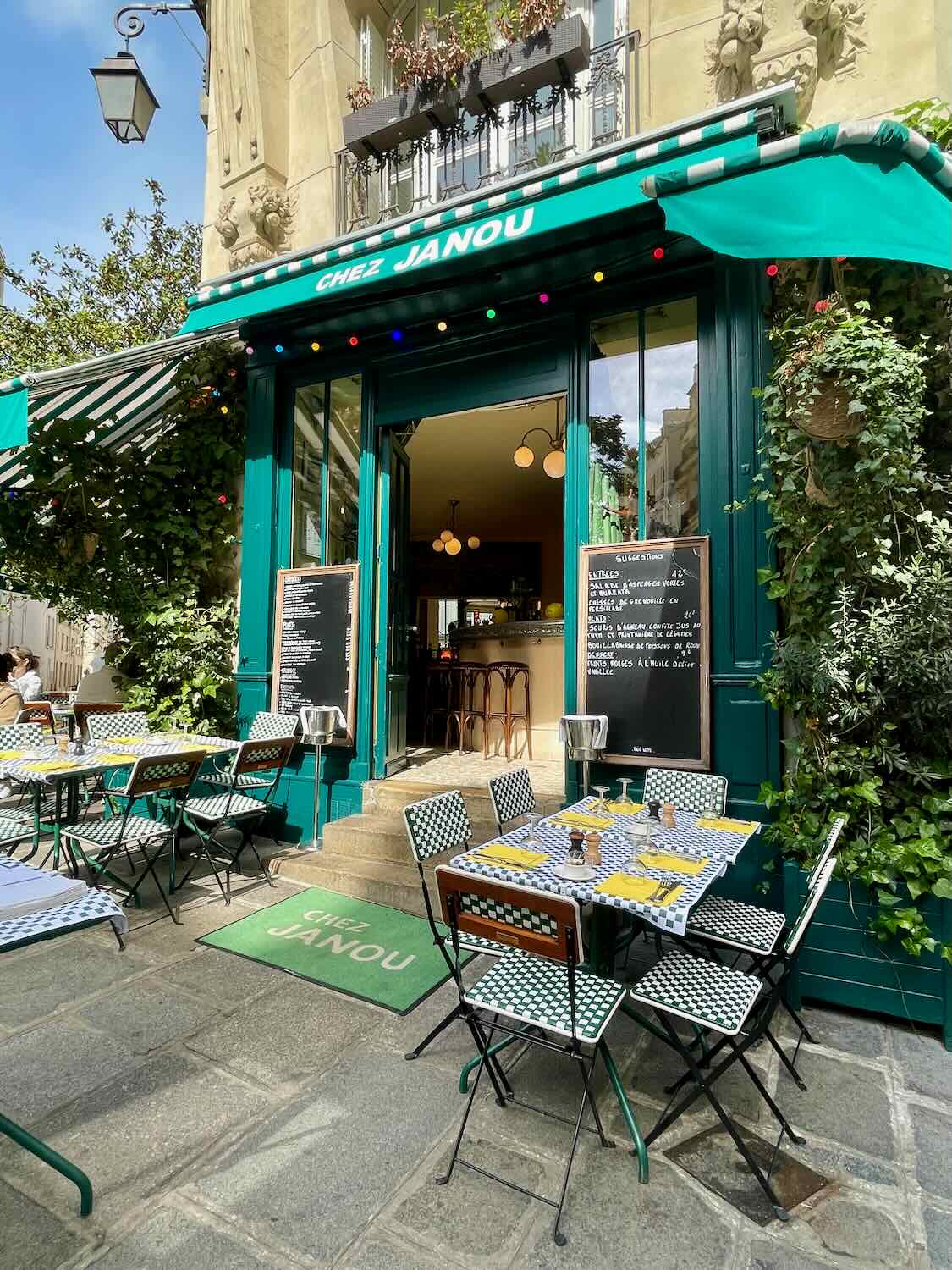 The inviting exterior of Chez Janou, a quaint Parisian bistro with a vibrant green facade and an outdoor seating area covered by a striped awning, set against a backdrop of traditional Paris architecture.
