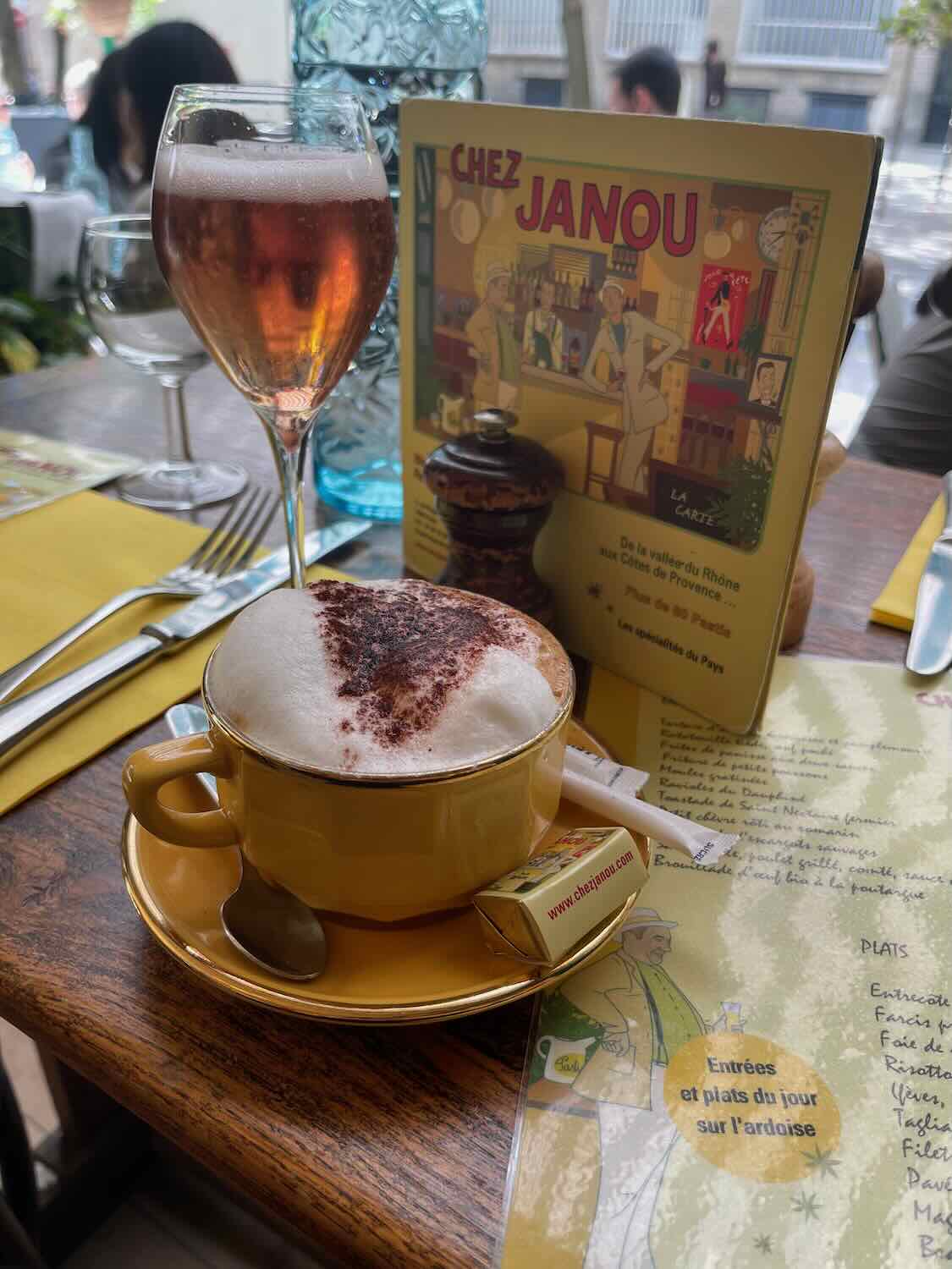 A table setting at Chez Janou featuring a cappuccino with a heart-shaped design in the foam and a glass of rosé, accompanied by a colorful menu, creating a cozy and welcoming dining experience.