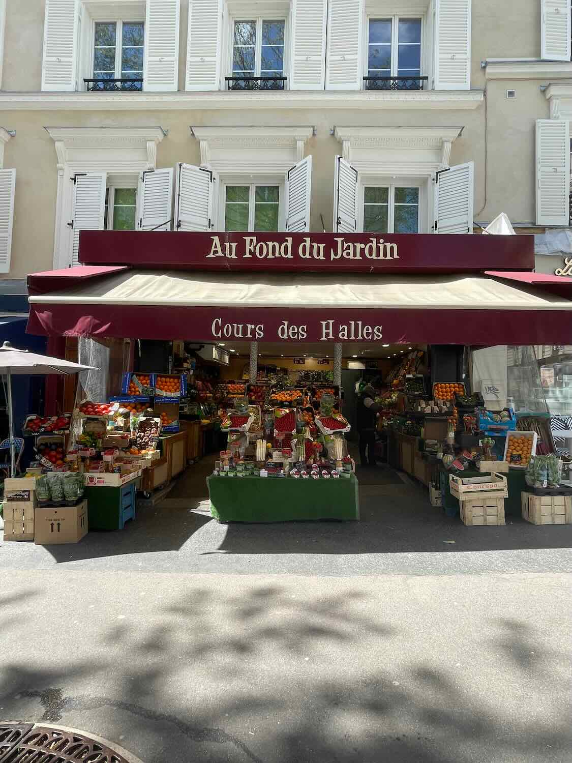 A vibrant market stall at Au Fond du Jardin in Paris, displaying a colorful assortment of fresh fruits and vegetables under a maroon awning on a sunny day.