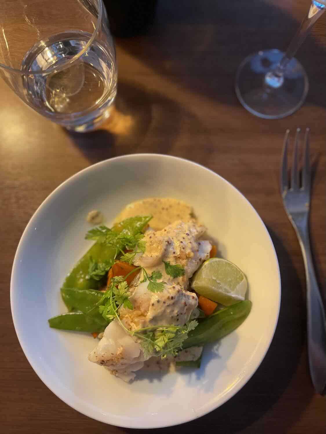 A close-up of a beautifully plated dish in Paris, featuring fish with vegetables, garnished with fresh herbs and a slice of lime, with a glass of water and wine in the background.