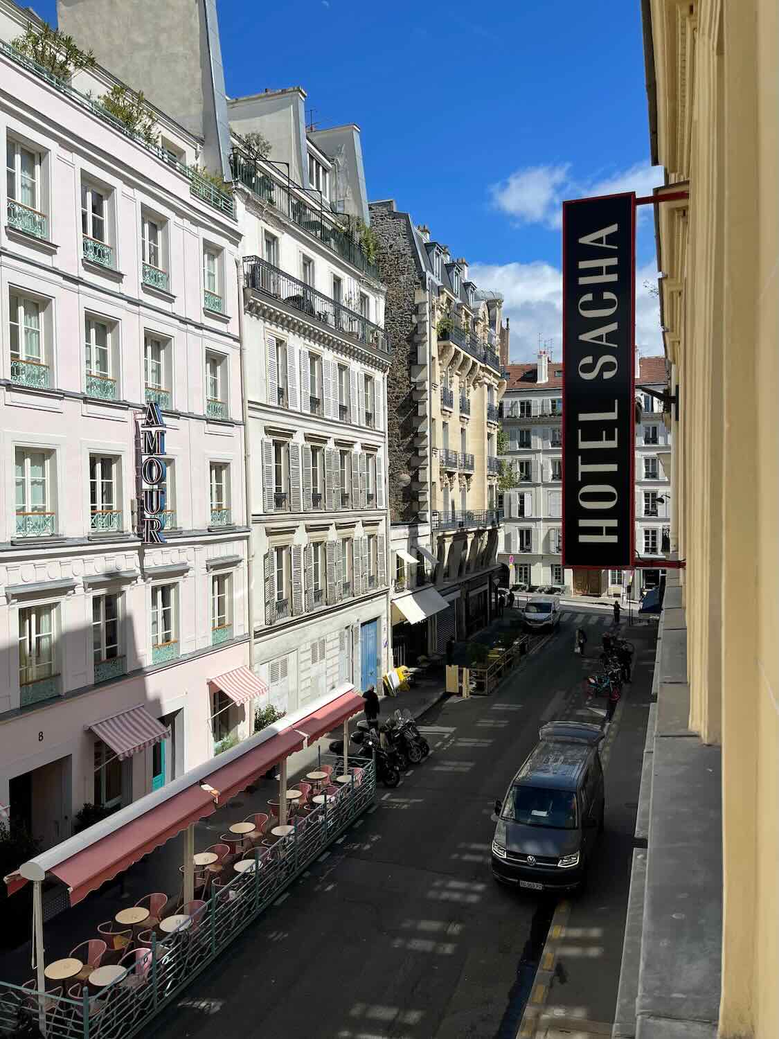 A picturesque view of the street outside Hotel Sacha in Paris, showing the charming facade of the hotel and nearby buildings with outdoor seating areas and a clear blue sky overhead.