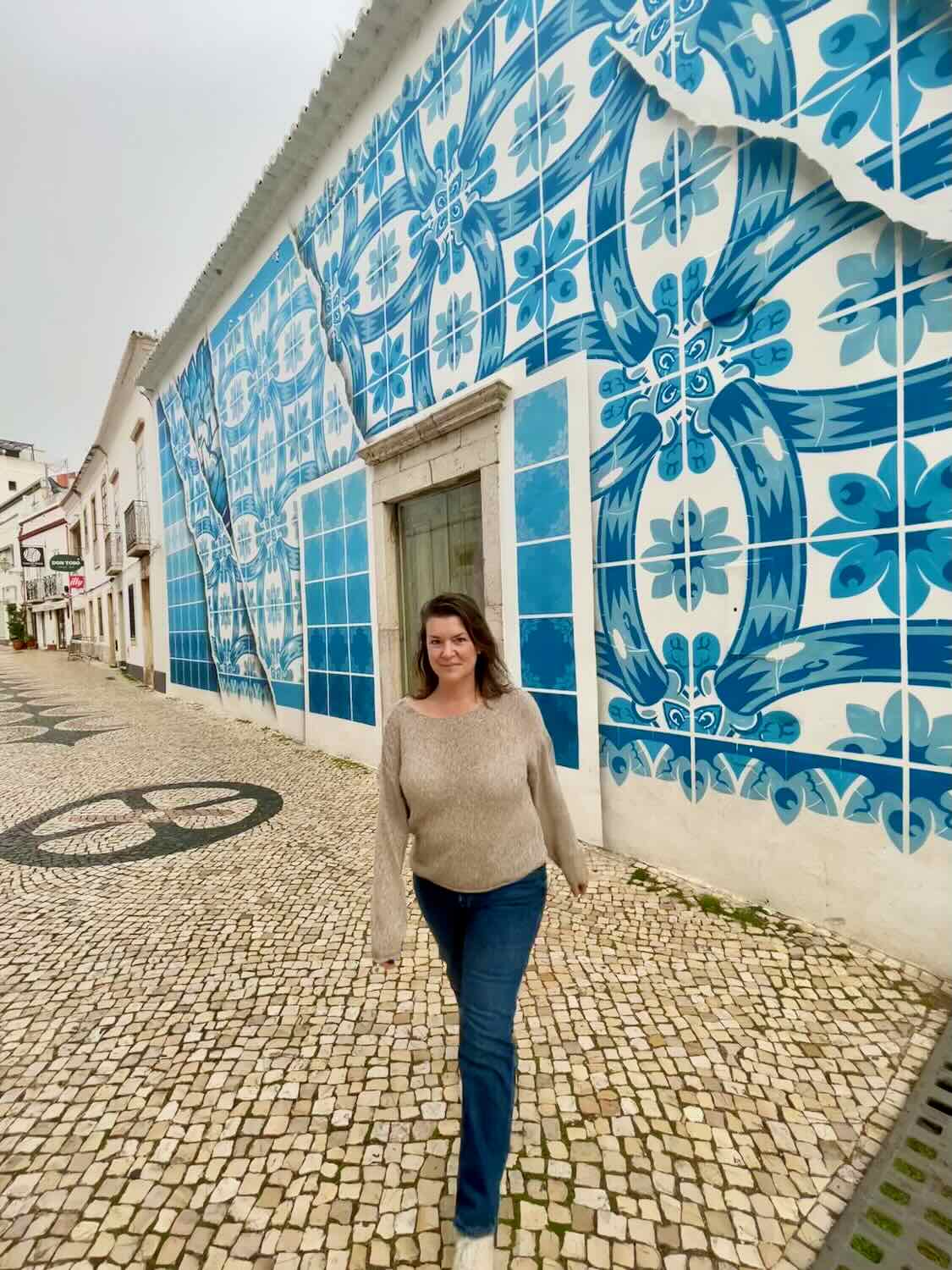 A woman walking on a cobblestone street in front of a building adorned with intricate blue and white tile designs in Portugal. She is wearing a beige sweater and blue jeans, with a relaxed and happy expression