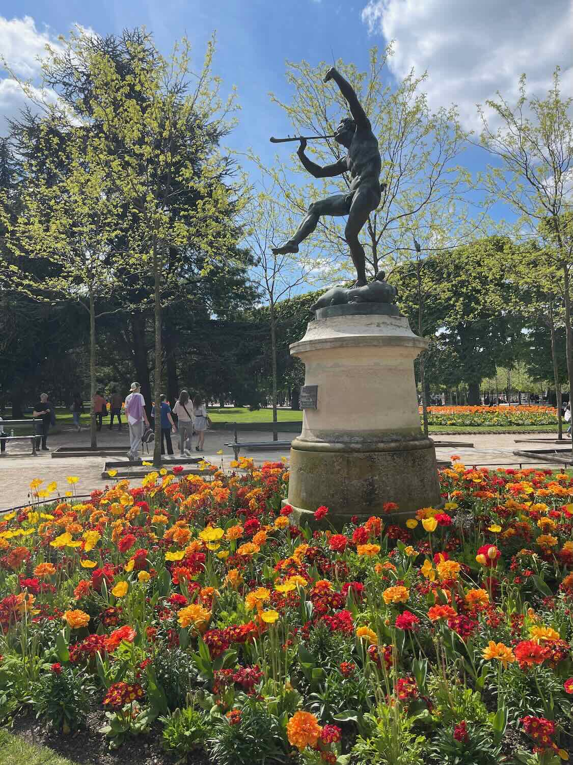 the famous Jardin du Luxembourg in Paris, featuring a bronze statue of a hunter surrounded by vibrant flowerbeds of red and orange marigolds under a sunny sky.