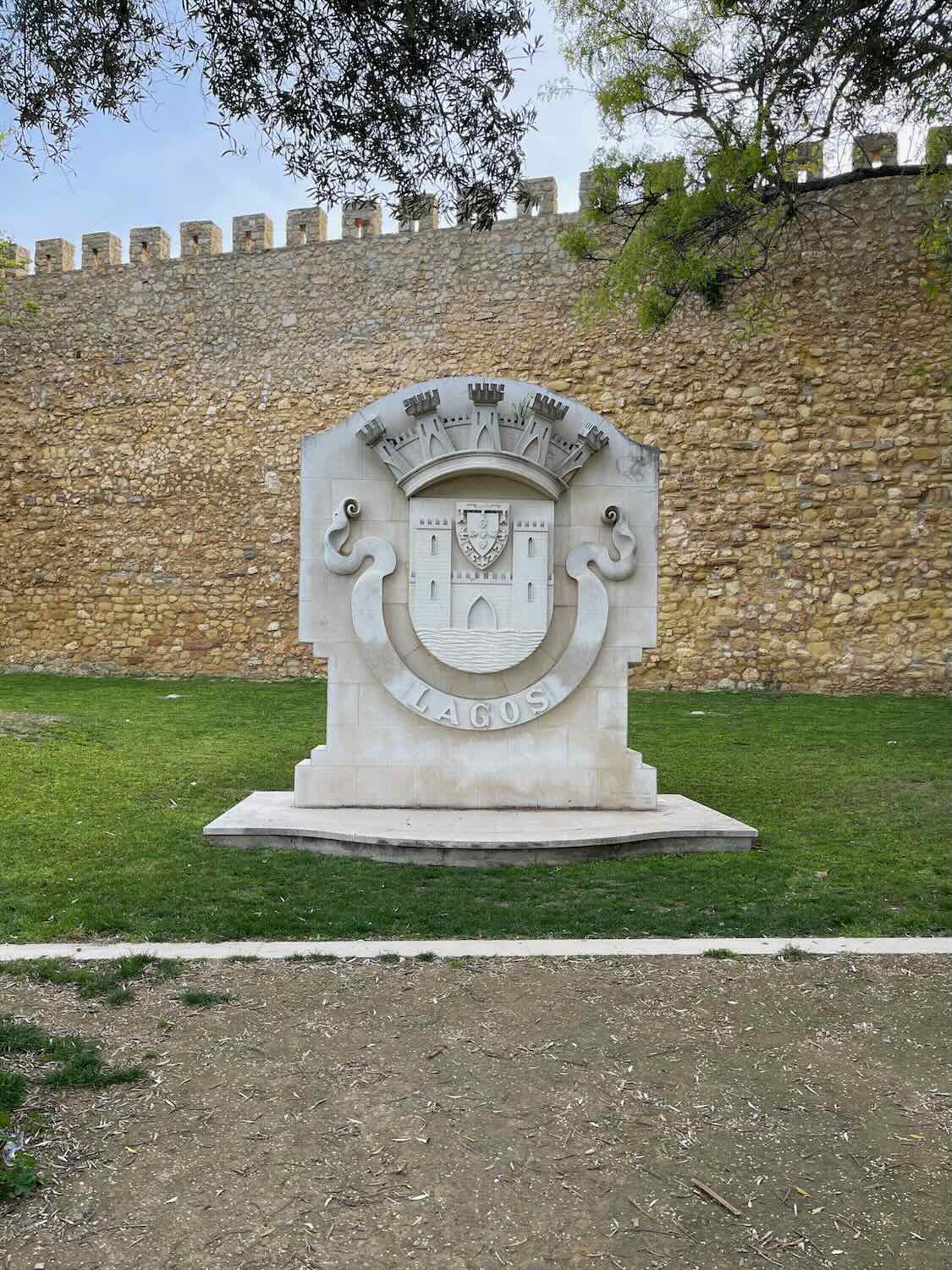 A large stone emblem featuring the word "Lagos" and a crest with a castle. The emblem is set against a backdrop of an ancient stone wall and surrounded by green grass.
