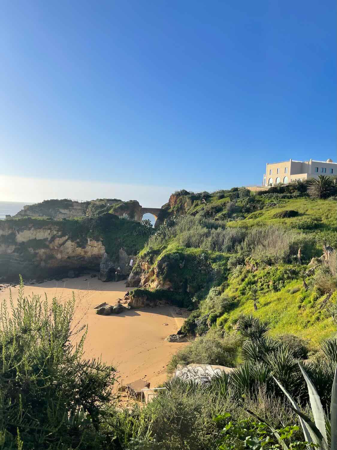 A lush green hill overlooking a secluded sandy beach with rocky cliffs and a stone arch bridge. A white building sits atop the hill, and the sky is clear and blue.