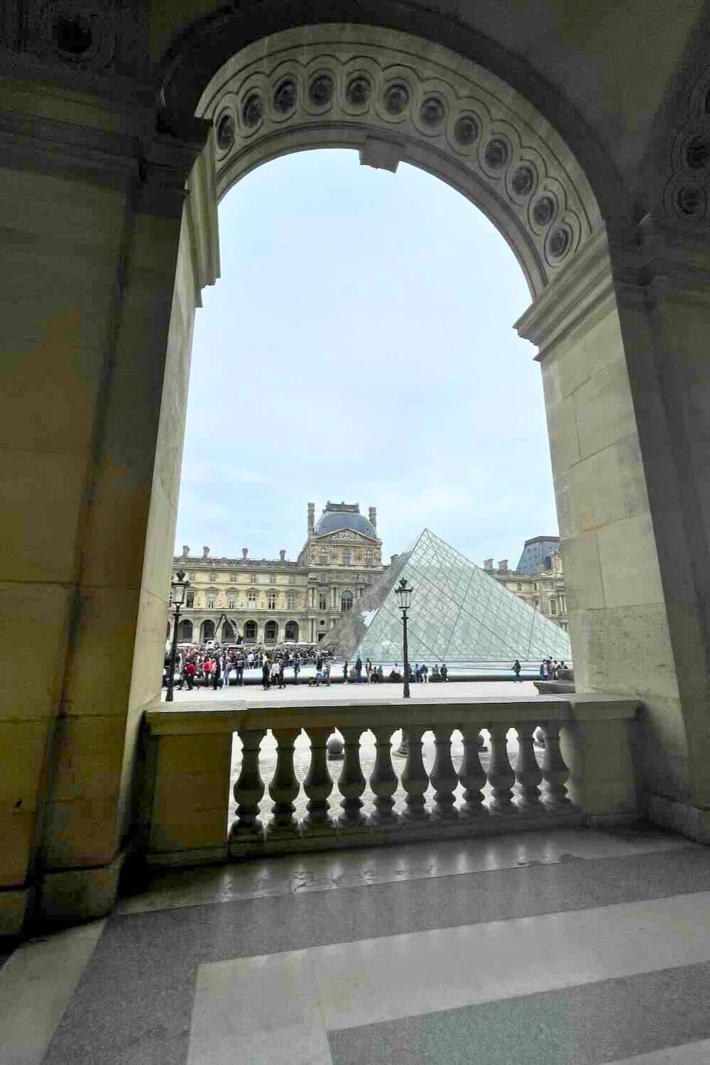 view from inside the Louvre Museum, looking out through an arched doorway towards the iconic glass pyramid and the museum's facade.