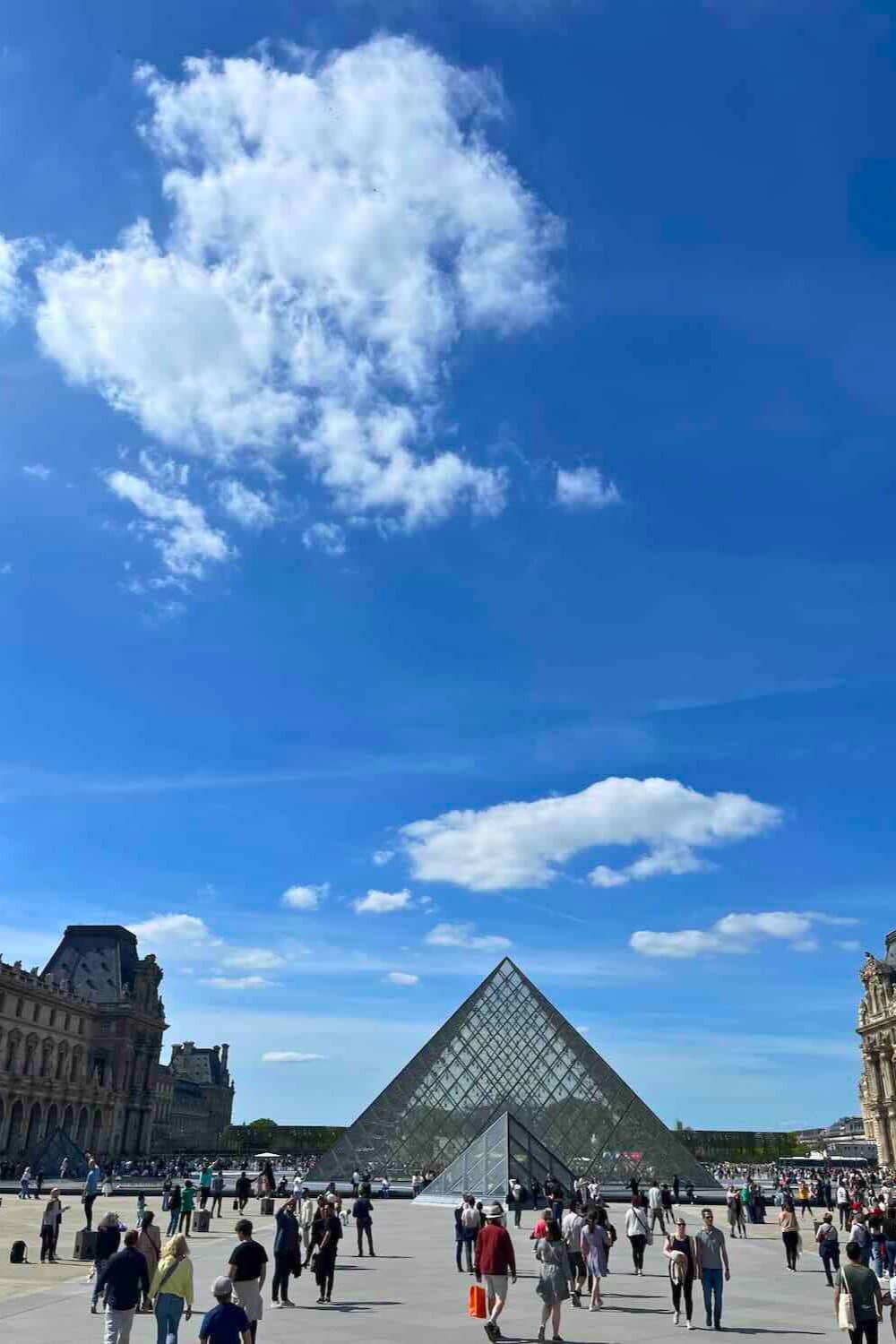 clear day scene at the Louvre Museum's courtyard, showcasing the large glass pyramid under a blue sky scattered with clouds.