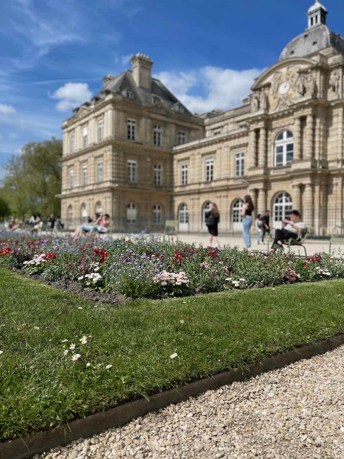 historical building within the Luxembourg Gardens, viewed across neatly trimmed lawns and colorful flowerbeds, under a clear blue sky.