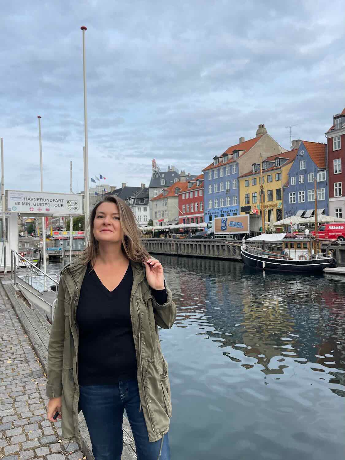 A woman standing in Nyhavn Harbor with colorful buildings and boats in the background.