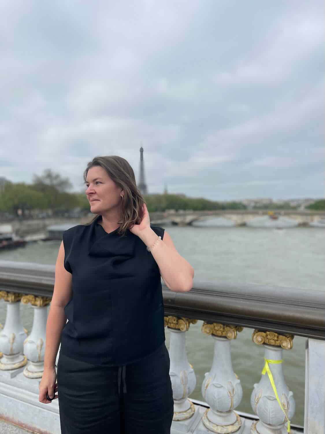 A solo female traveler standing by the Seine river in Paris, looking to the side with the Eiffel Tower in the distant background. She is dressed in a black sleeveless top and black pants.