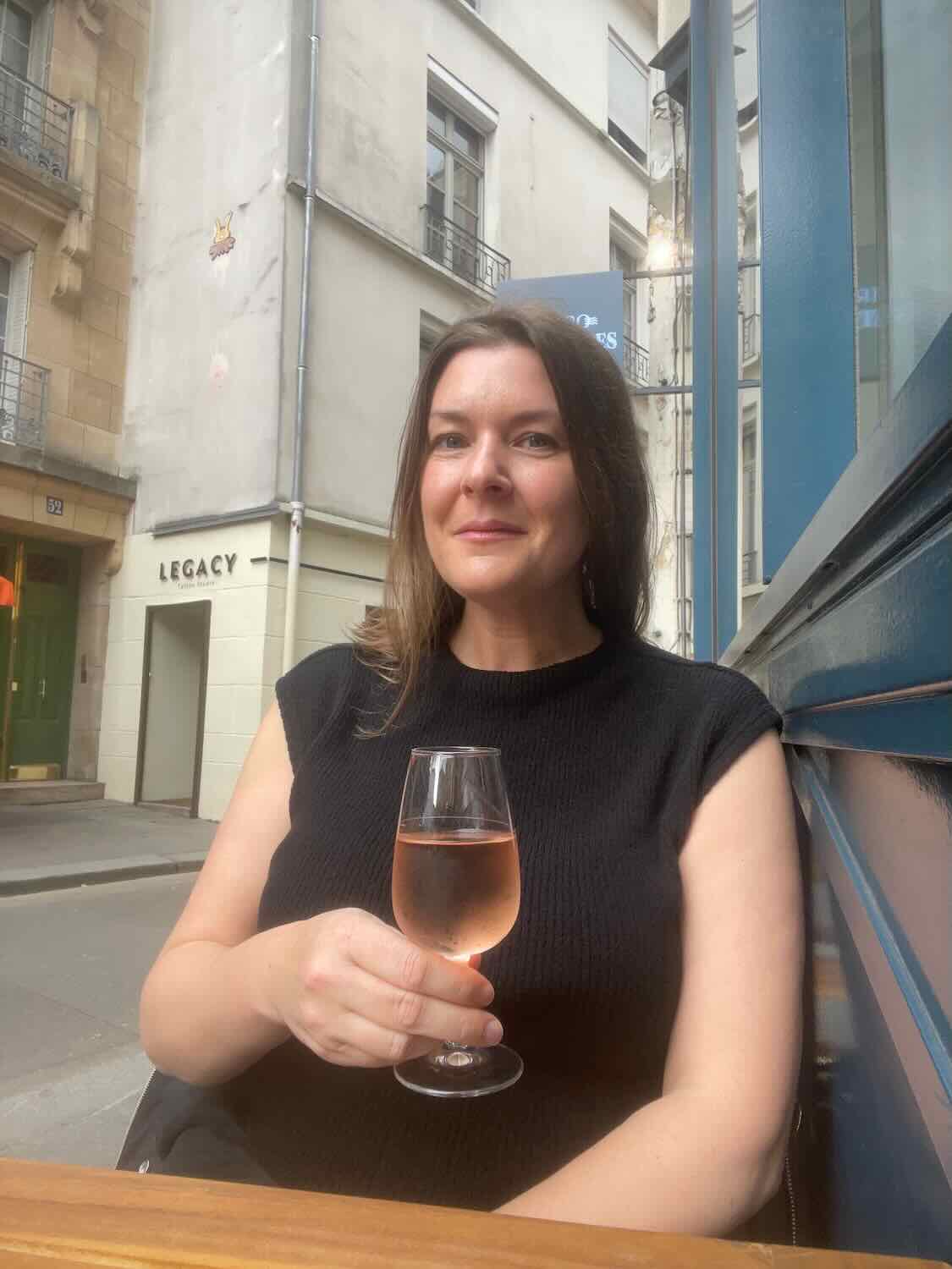 A woman sitting outside a cafe in Paris, holding a glass of rosé wine and smiling at the camera. The street and building facades can be seen in the background.