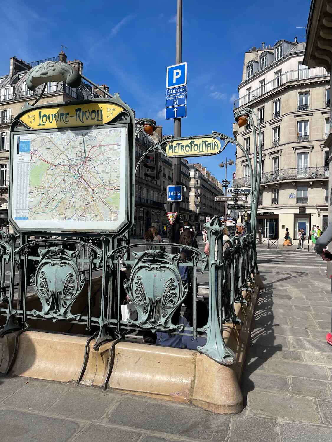 The image features the Louvre-Rivoli metro station entrance in Paris, characterized by its iconic Art Nouveau design. The green, ornate ironwork frames a city map and the metro sign. The backdrop shows typical Parisian architecture and bustling city life with pedestrians in the area.