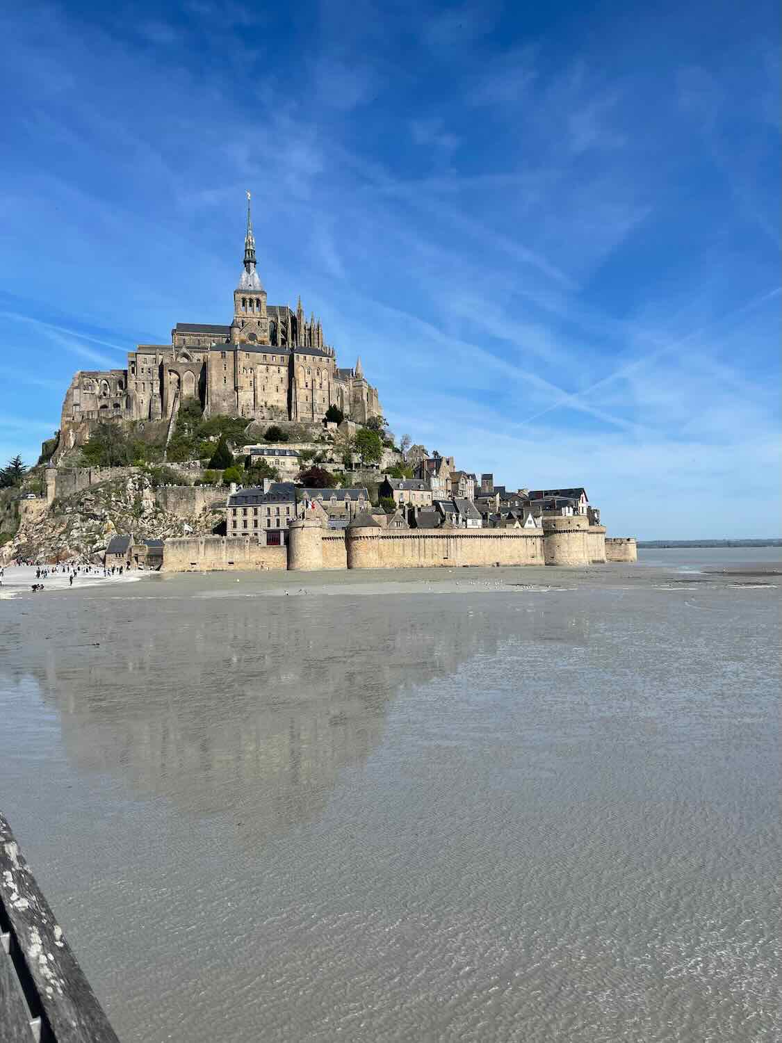 Clear day view of Mont Saint Michel, showing the entire island and abbey against a bright blue sky, reflecting on a shallow bay.