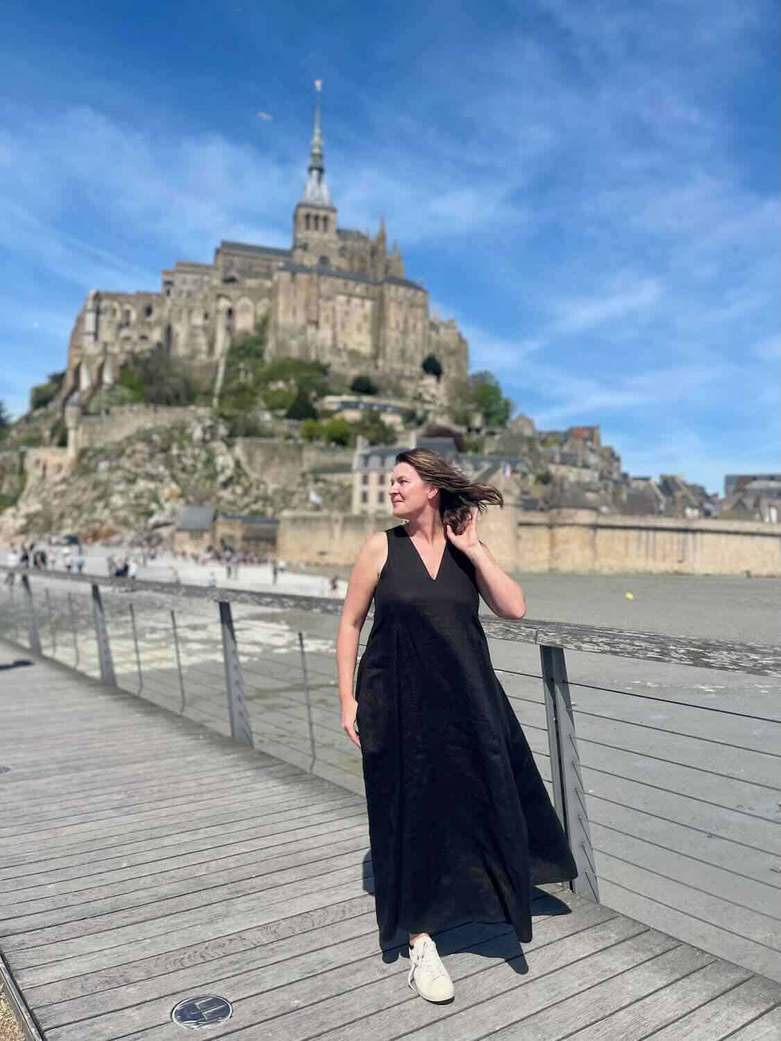 Solo female traveler standing on a wooden boardwalk with Mont Saint Michel in the background, highlighting the personal experience of visiting.