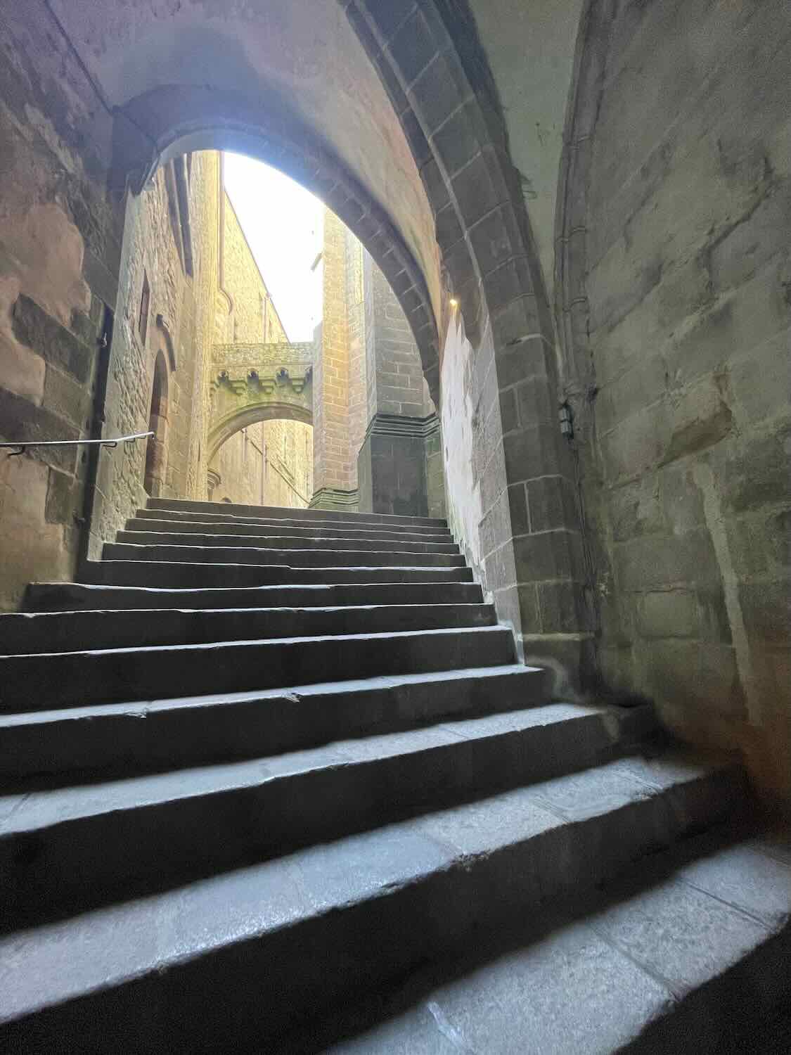 Atmospheric shot inside Mont Saint Michel showing a stone staircase leading upwards, framed by arches and illuminated by soft natural light.