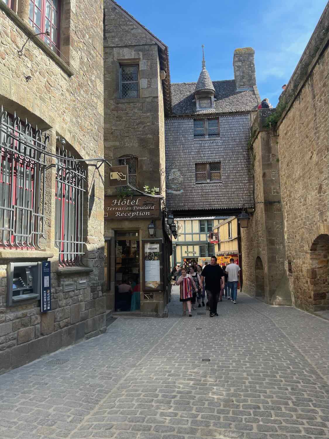 Historic stone buildings line a bustling narrow street in Mont Saint Michel, complete with quaint shop fronts and cobblestones, under a clear blue sky.