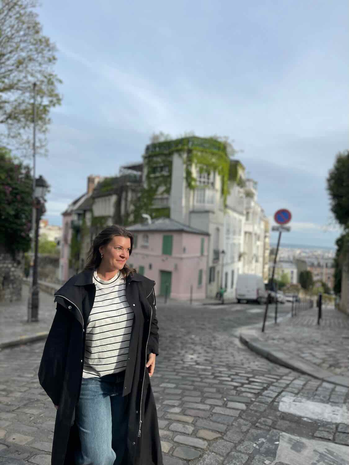  a woman walking on a quaint cobblestone street in the Montmartre neighborhood. The street is flanked by charming, pastel-colored buildings covered in greenery. The woman is dressed casually in jeans and a striped top, with a long black coat, and appears relaxed and content, embodying the solo traveler exploring Paris.