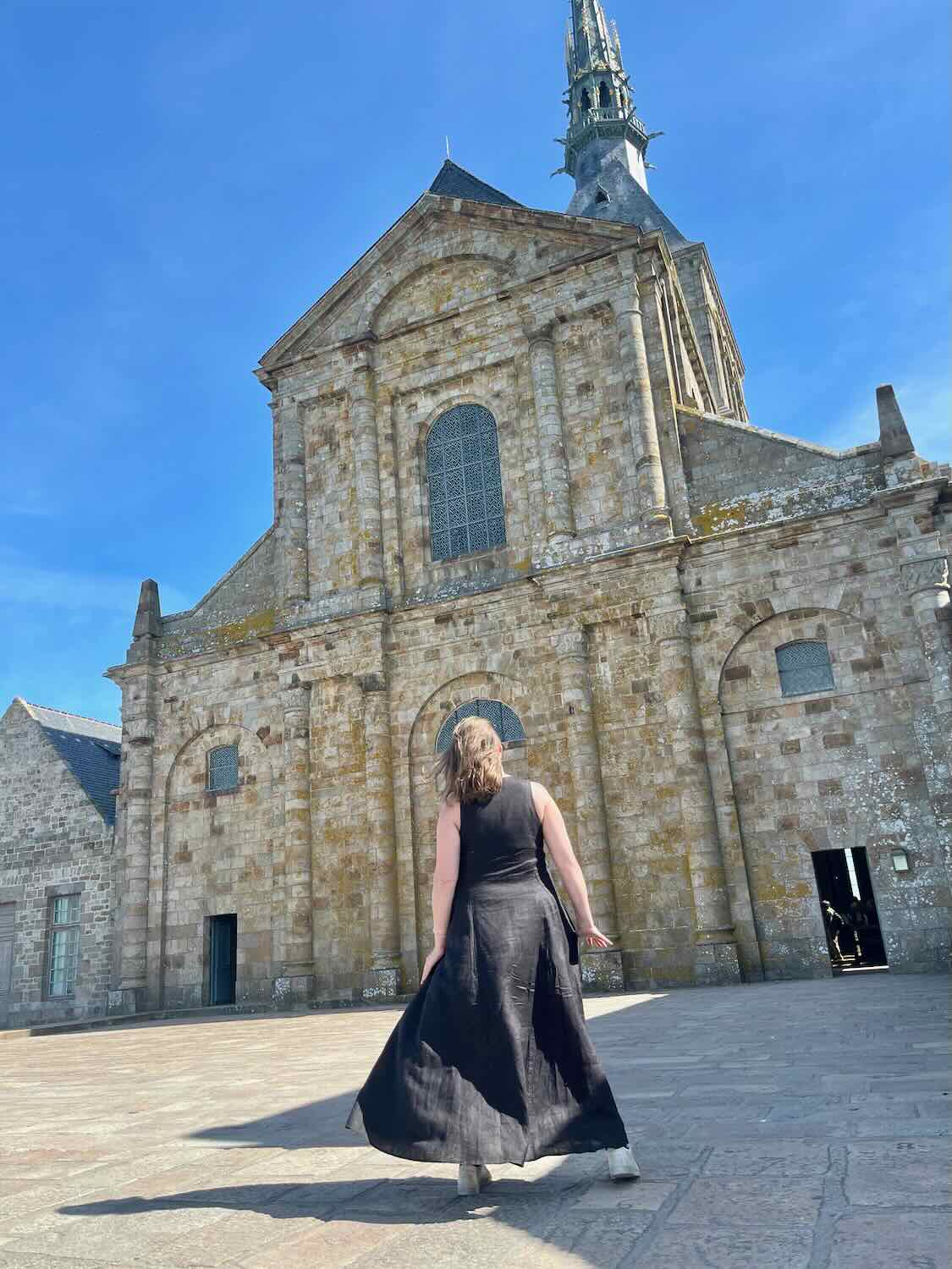 A woman in a black dress walks towards the imposing stone facade of Mont Saint-Michel Abbey under a clear blue sky.