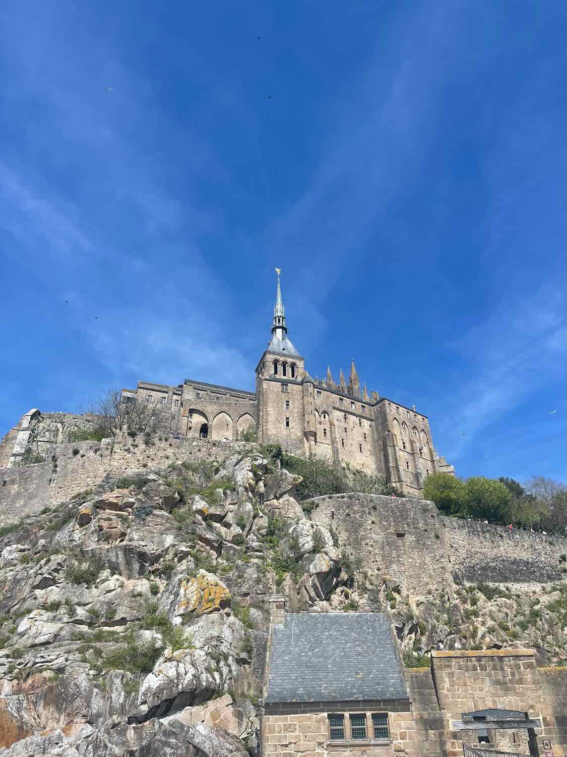 The spire of Mont Saint Michel Abbey rises high above rocky terrain and medieval structures, framed by a clear blue sky.