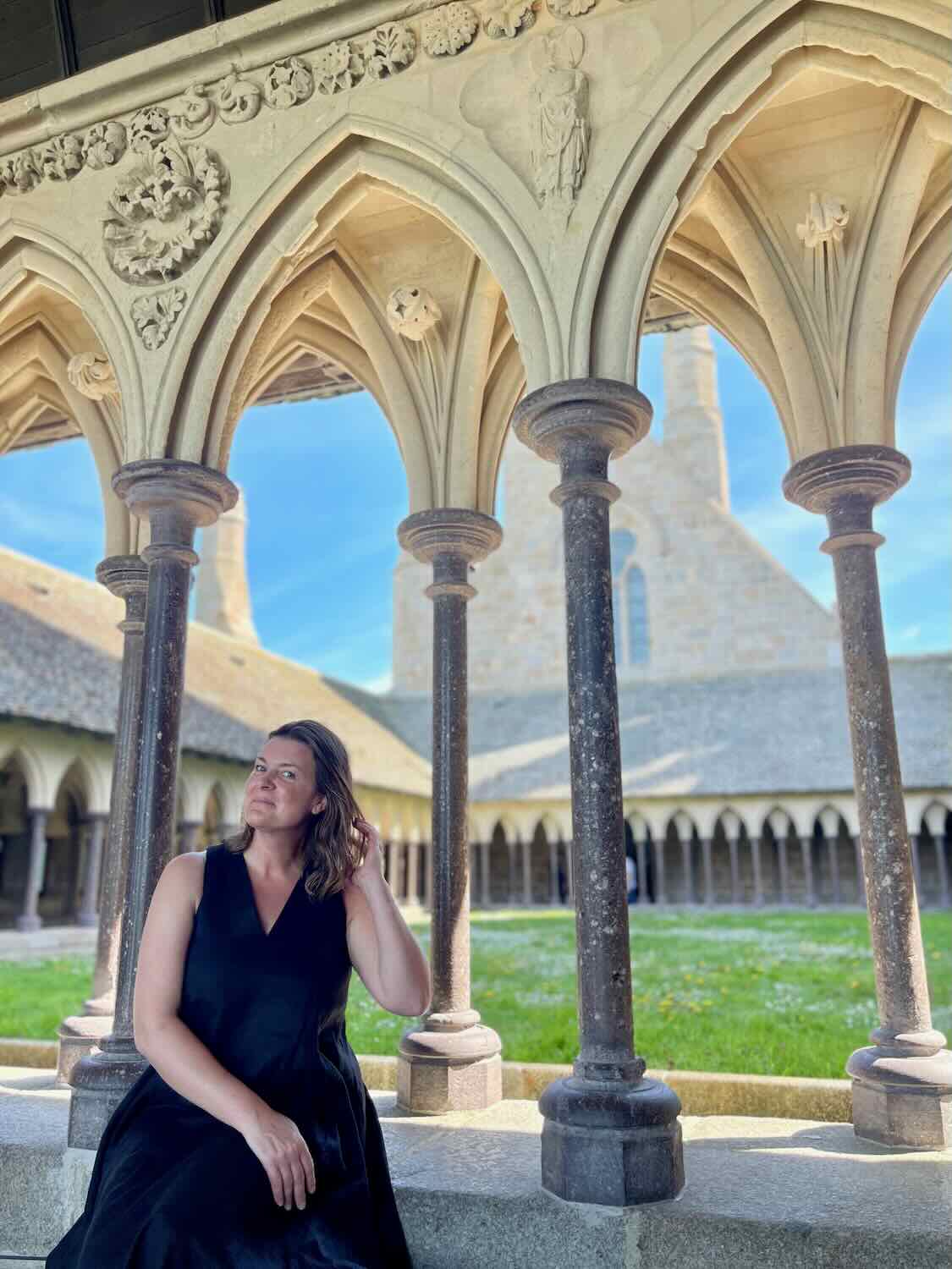 A solo female traveler sits thoughtfully in the Gothic cloisters of Mont Saint Michel Abbey, surrounded by ornate arches and a serene atmosphere.
