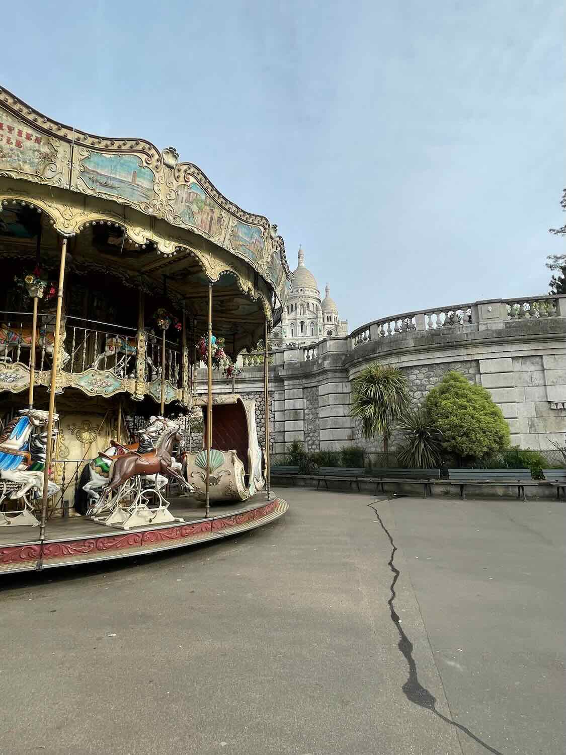 The image features a charming old carousel with intricate designs and classic painted horses, located near the base of the Sacré-Cœur Basilica in Montmartre, Paris. The scene is picturesque, blending the whimsical nature of the carousel with the grandeur of the historic basilica in the background, under a slightly cloudy sky.