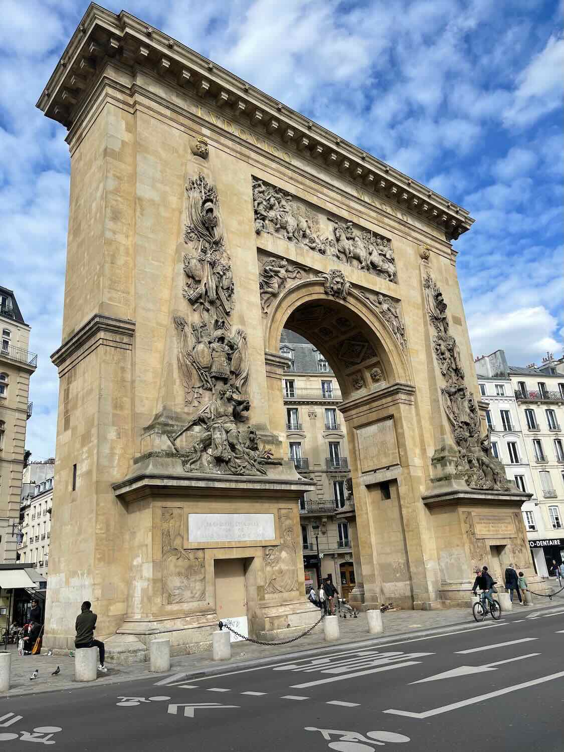 The photo shows the Porte Saint-Denis, a large arch monument in Paris, France. It features intricate relief sculptures and is made of stone, standing prominently against a clear sky. The surrounding area includes pedestrians and cyclists, capturing a lively urban scene.