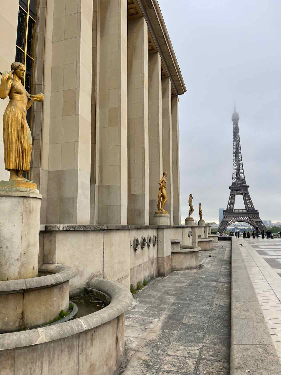 A view of the Trocadéro Gardens in Paris, featuring several golden statues along a grand building, with the Eiffel Tower rising in the misty background.