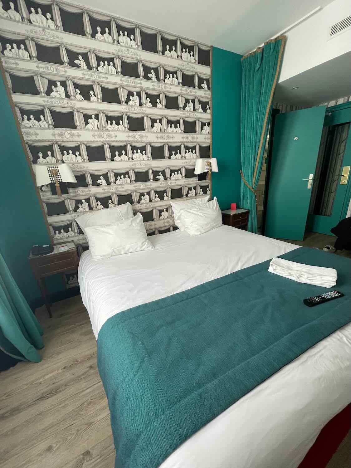 A stylish hotel room featuring a unique black and white sculpture-themed wallpaper, with neatly arranged white bedding on a bed, accented by a teal throw and matching teal curtains, offering a chic and artistic atmosphere.