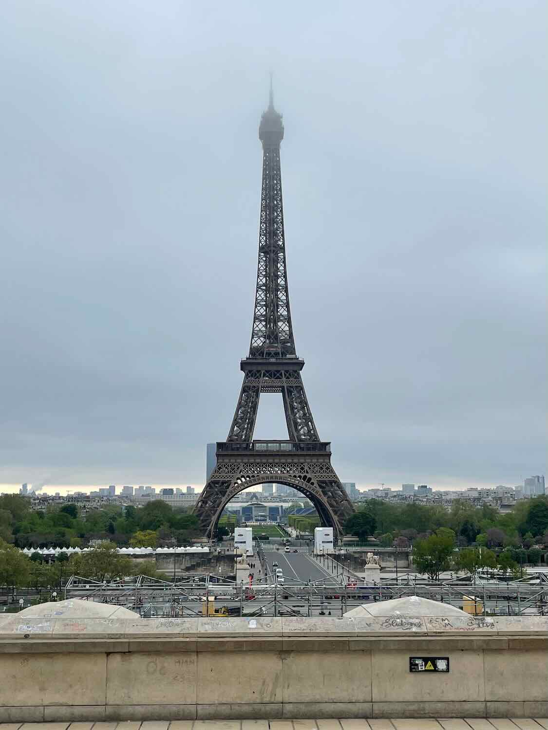 A view of the Eiffel Tower on a cloudy day, with its top partially obscured by mist. The foreground shows the plaza area with minimal foot traffic.