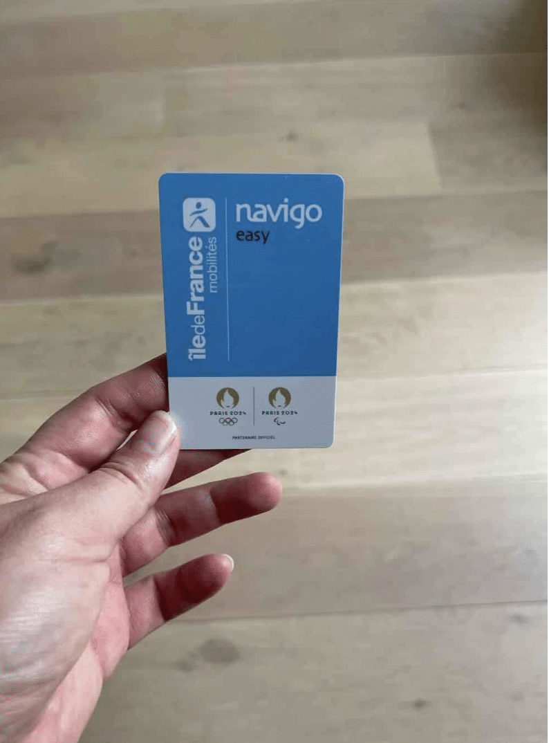 A close-up of a hand holding a Navigo Easy card, used for public transportation in Paris. The card is blue and white, featuring the Île-de-France Mobilités logo and the Paris 2024 Olympic and Paralympic logos