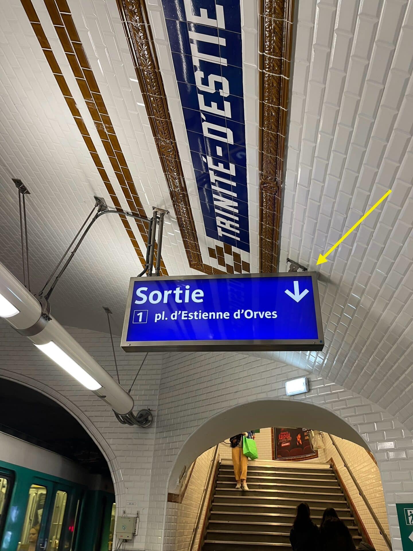 nside a Paris metro station, highlighting the iconic white tiled walls with a blue sign reading "TRINITE-D'ESTIENNE D'ORVES" and a digital blue sign pointing towards the exit at Place d'Estienne d'Orves.