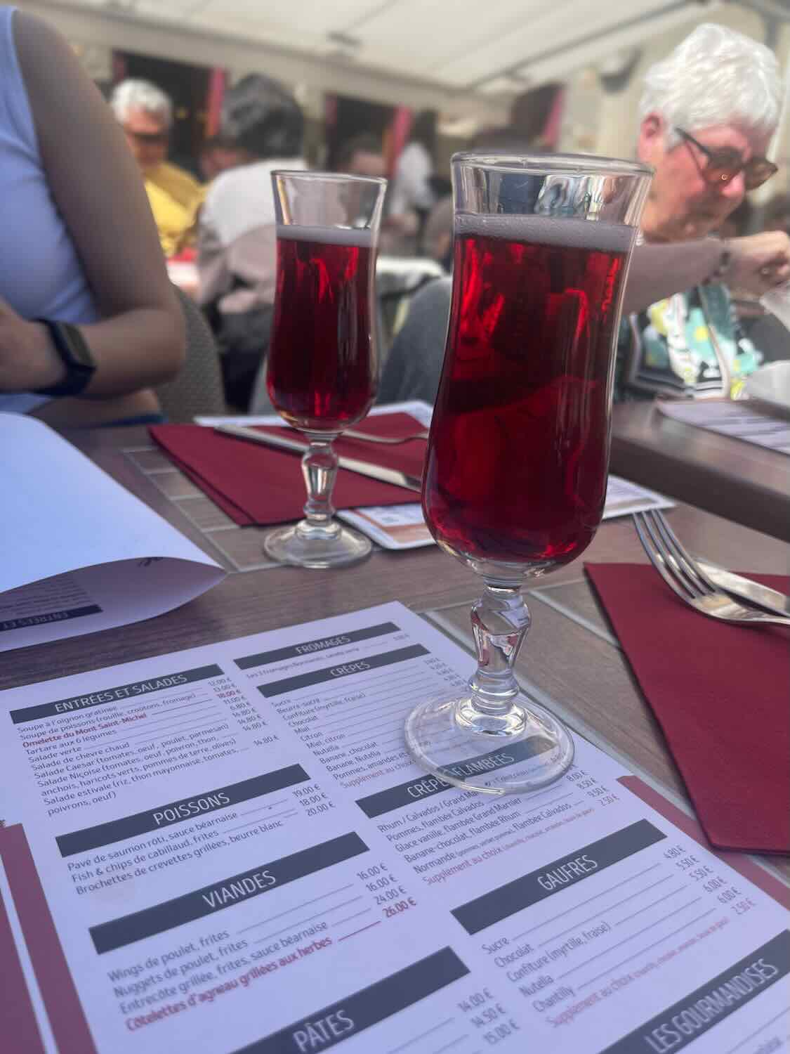 Two glasses of red kir on a restaurant table in Mont Saint Michel, with a blurred menu and diners in the background, offering a glimpse into local culinary experiences.