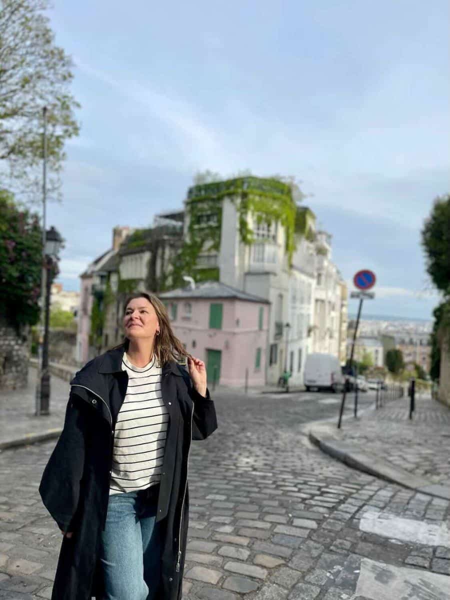A woman wearing a black coat and striped shirt walks on a cobblestone street in Paris. Behind her, there is a charming pink building with greenery covering parts of it. The sky is clear, suggesting a pleasant day.