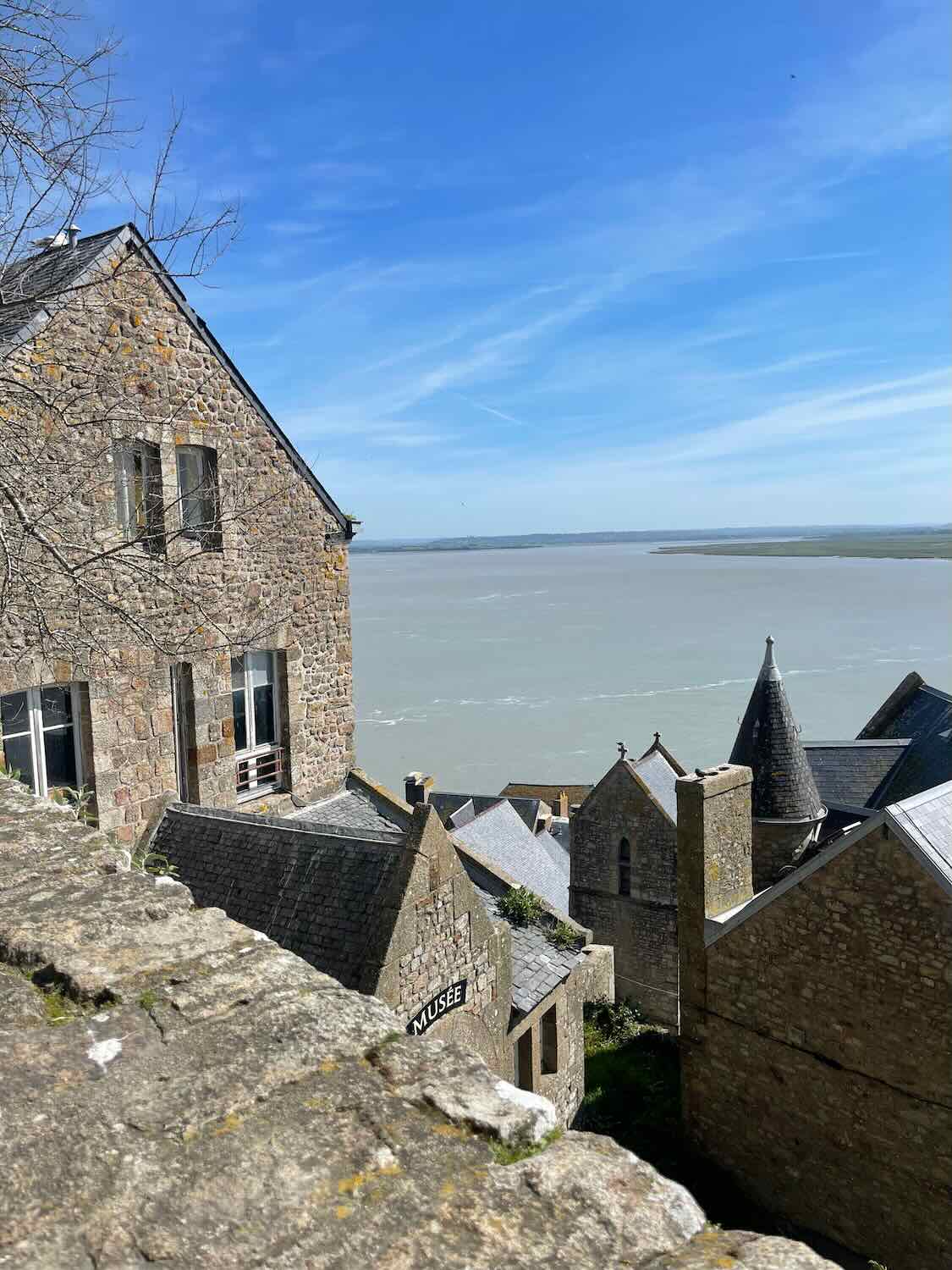 A picturesque view over the rooftops of Mont Saint Michel, looking out to the expansive bay under a clear sky, with historical buildings densely packed together.