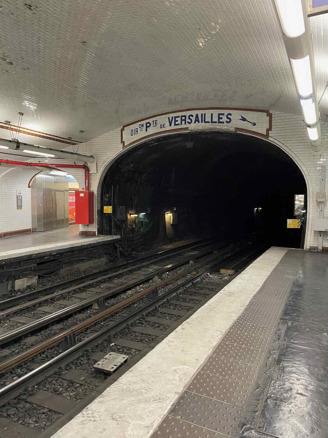 An empty platform at a Paris metro station with "Direction Pt de Versailles" sign, typical white metro tiles, and tracks leading into a dark tunnel, embodying the everyday commute in the city.