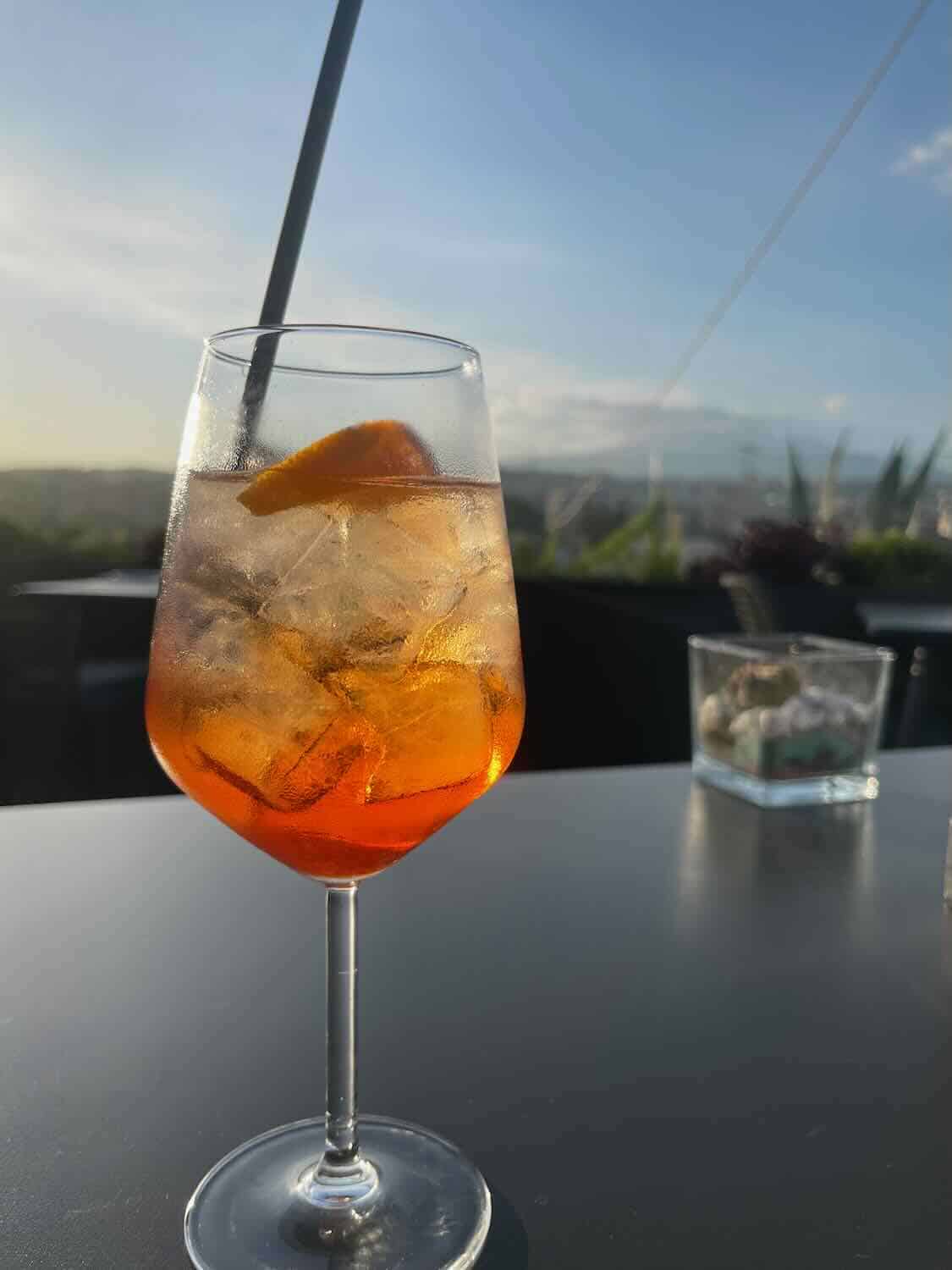 A photo of a glass of Aperol Spritz with ice and an orange slice, sitting on a table with a straw. The background shows an outdoor setting with a clear sky and distant hills, creating a relaxed atmosphere.