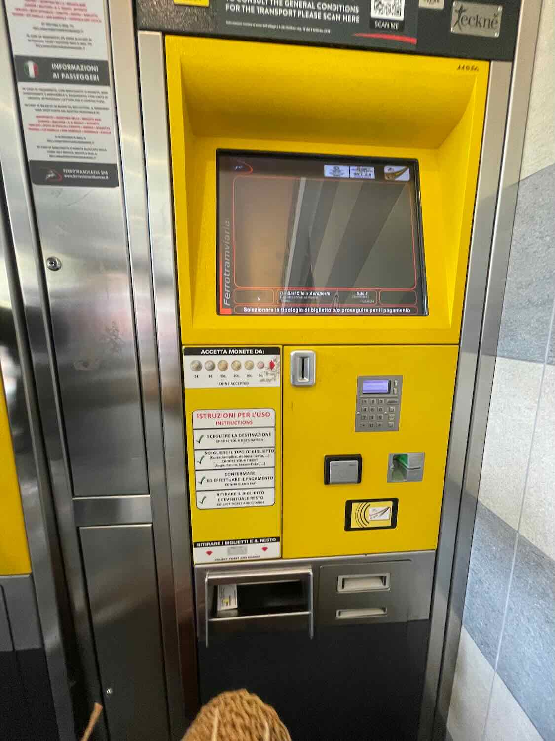 A yellow ticket machine at Bari airport, with instructions in Italian and options for selecting destinations and ticket types. The screen is currently off, and the machine accepts both coins and cards for payment.