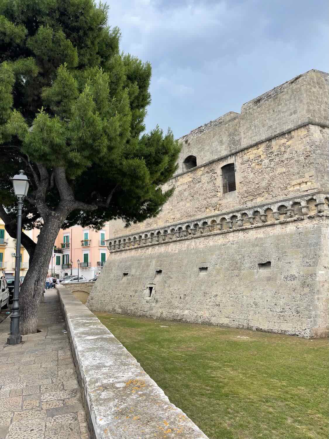 A photo of the exterior of a historic castle in Bari, Italy. The stone structure features thick walls with small windows and a crenelated parapet. A large tree and a stone pathway with a lamp post are visible in the foreground, and colorful buildings can be seen in the background.