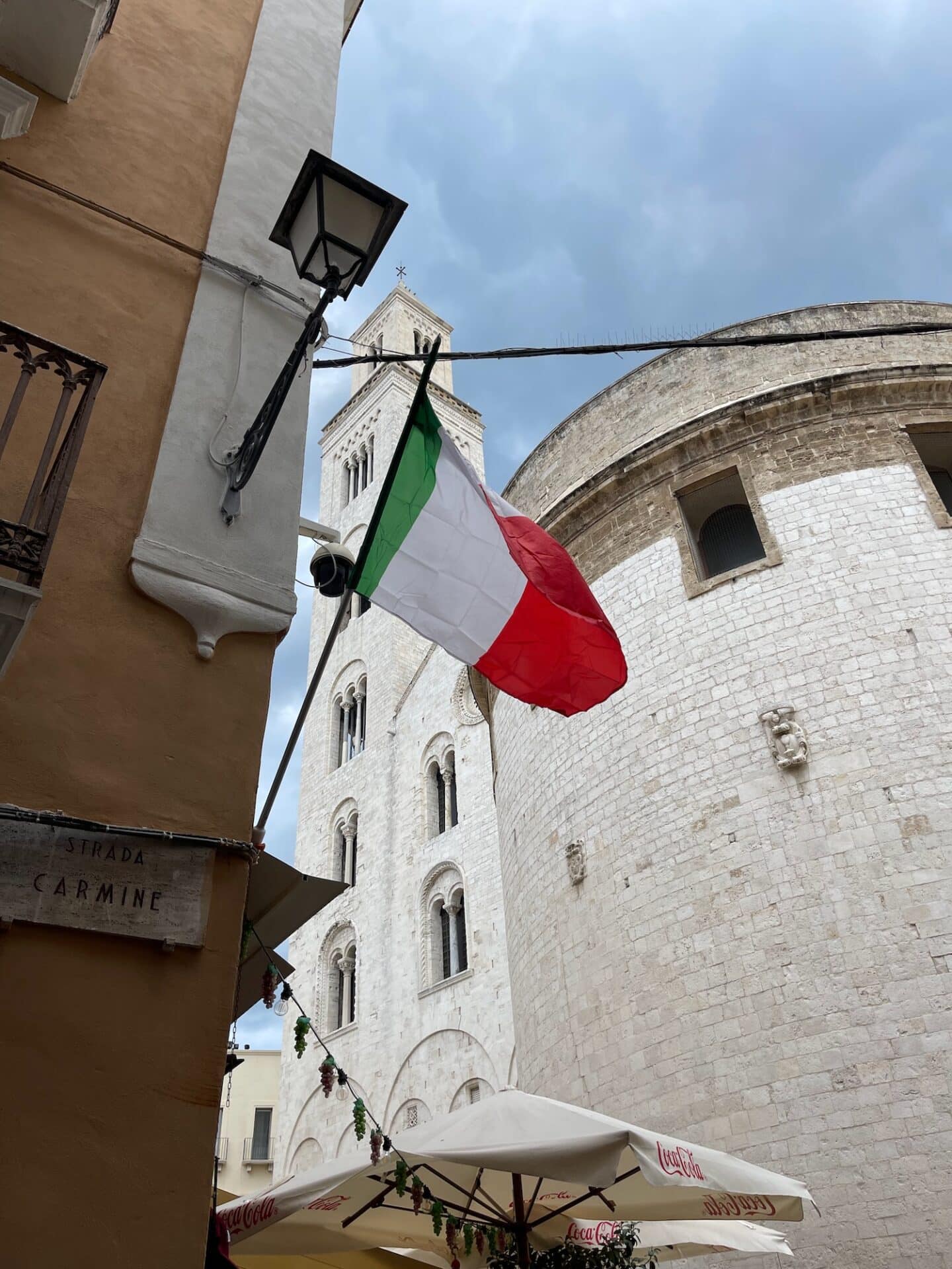 An Italian flag waves in the wind with the Basilica of San Nicola in the background. The basilica's white stone facade and tower are visible against a cloudy sky. A street sign for 'Strada Carmine' is also seen on the wall.