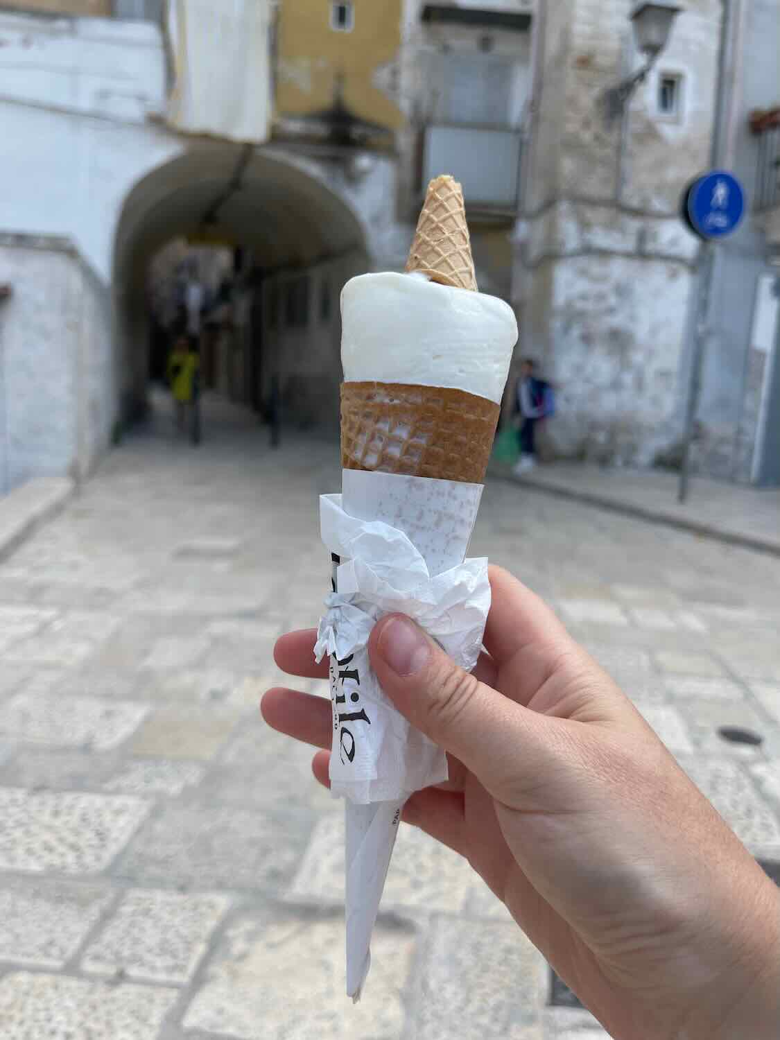 A close-up photo of a hand holding a gelato cone with a scoop of white gelato in Bari, Italy. The background features a narrow street with historic buildings and an archway, providing a charming and picturesque setting.