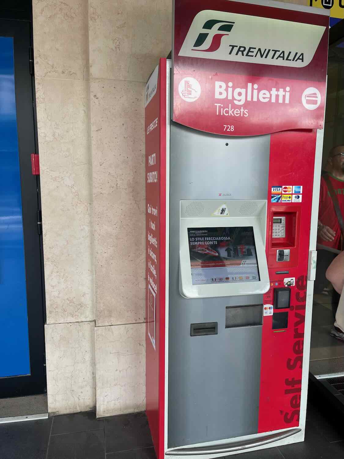 A photo of a Trenitalia ticket machine in Bari, Italy. The machine is red and grey with a touchscreen interface and various payment options displayed. It is located against a stone wall, with signage indicating 'Biglietti' (Tickets) and 'Self Service'.